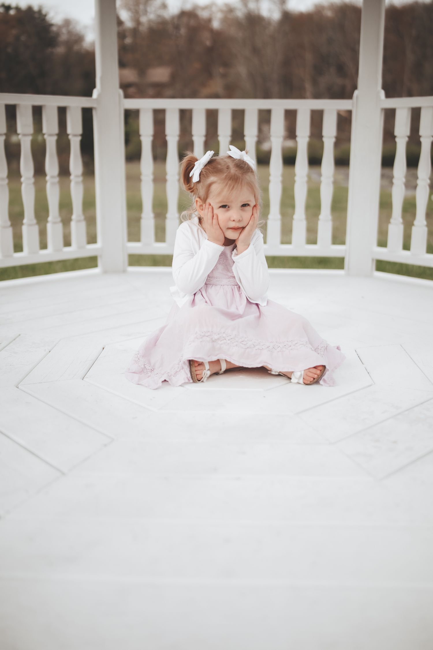 A child in a white dress lies on a white wooden deck with white railings and autumn trees in the background.