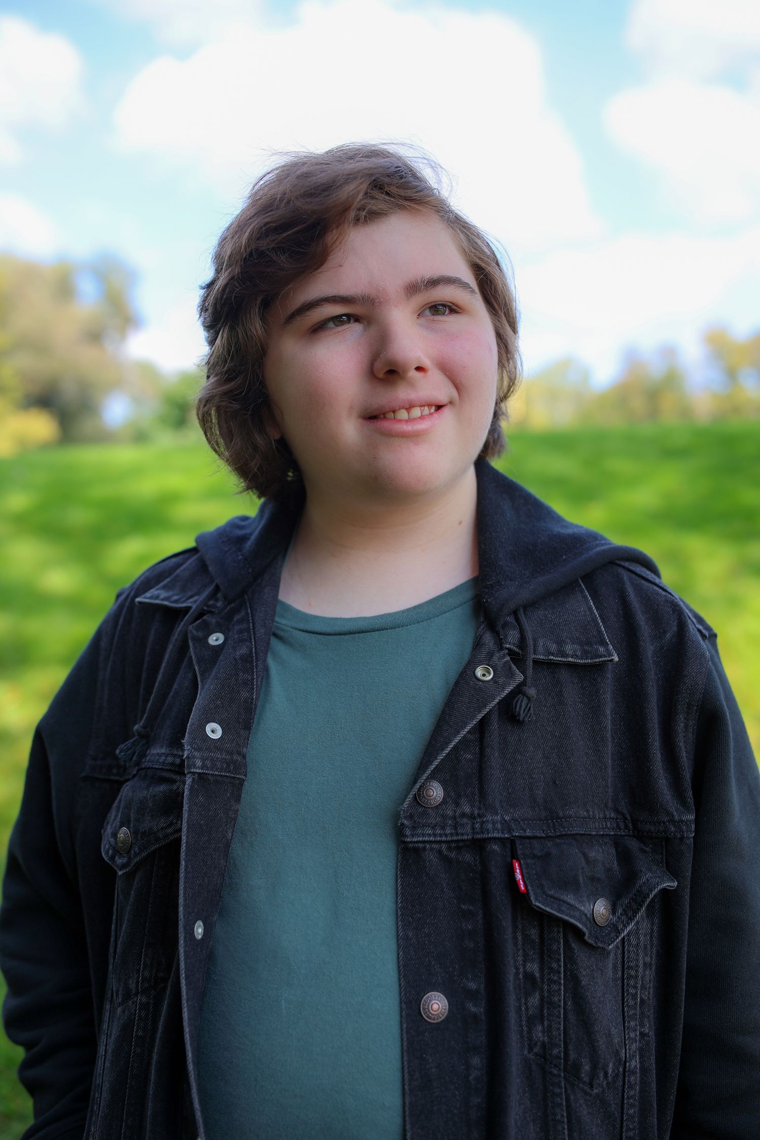 Person wearing a black denim jacket and green shirt poses outdoors against a sunny blue sky background.