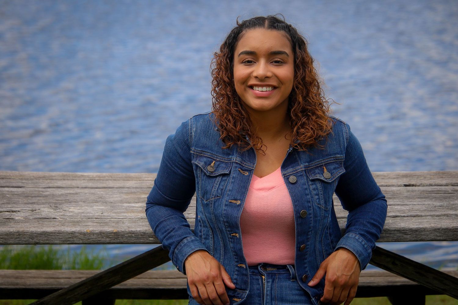 A person in a denim jacket sits outdoors on a wooden dock near a lake, smiling warmly at the camera.