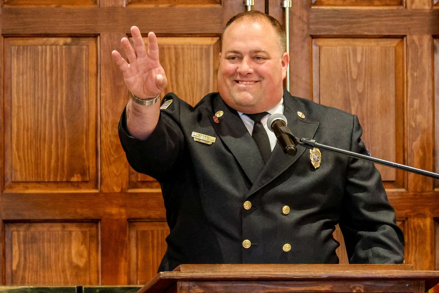 A person in a dark uniform waves while standing at a wooden podium in front of wood-paneled walls.
