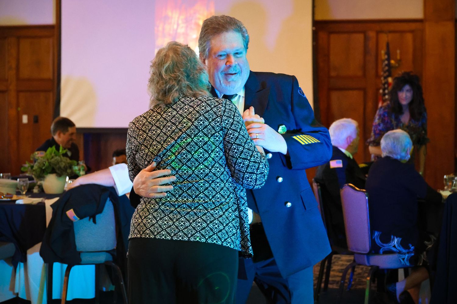 Two people dance together in a wood-paneled banquet hall with blue lighting and other guests seated at tables.