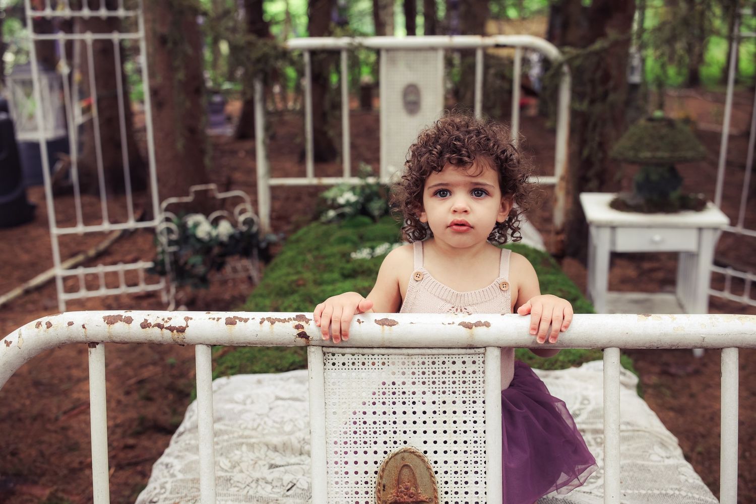 A toddler peeks over a white lattice fence in a garden setting with brick buildings in the background.