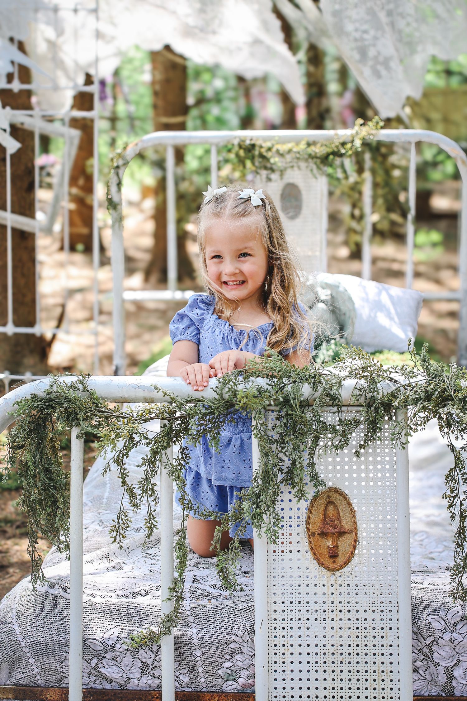 Vintage white iron bed decorated with greenery and lace in an outdoor garden setting.