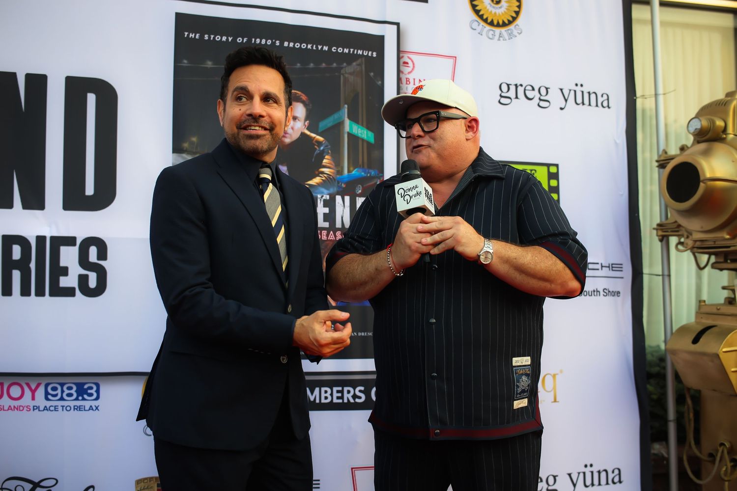 Two people in casual and formal attire share drinks while standing at an event with promotional signage in the background.