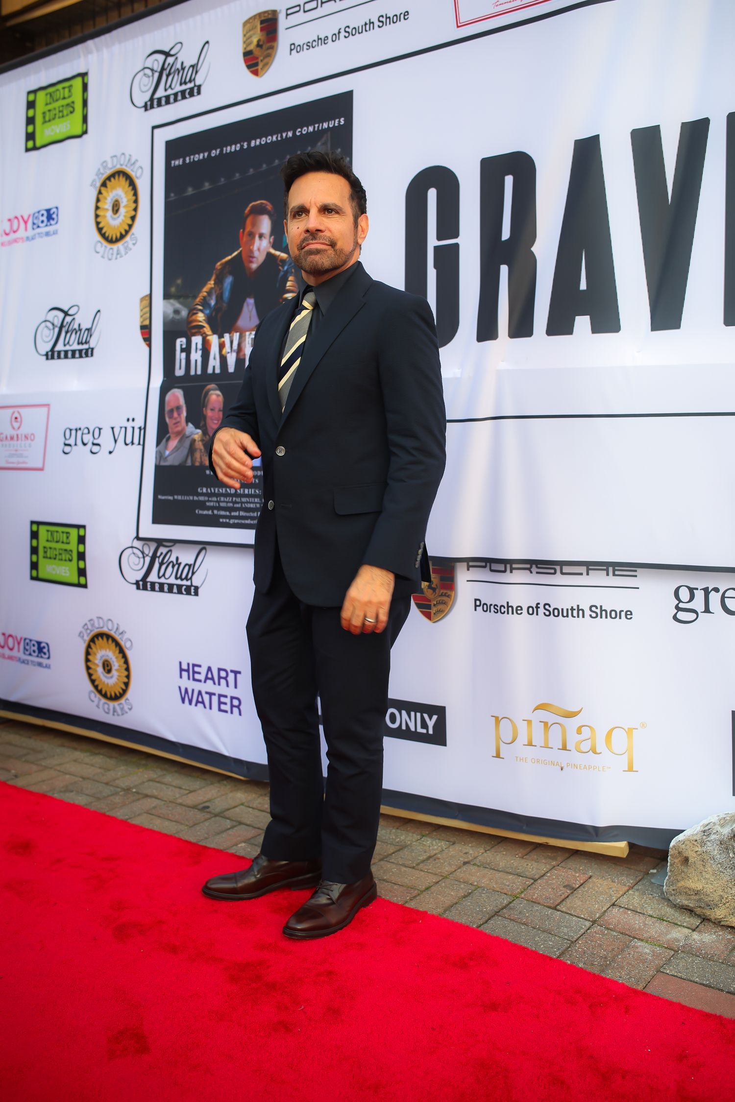 Black dress shoes and suit pants visible on a red carpet with sponsor logos and banners in the background.