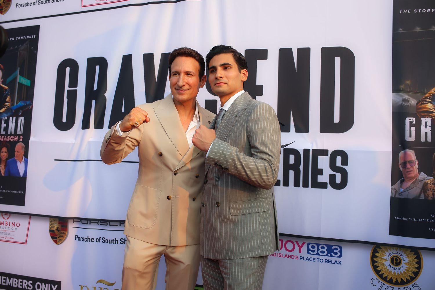 Two people in suits pose together at a movie premiere event with promotional posters and banners visible in background.