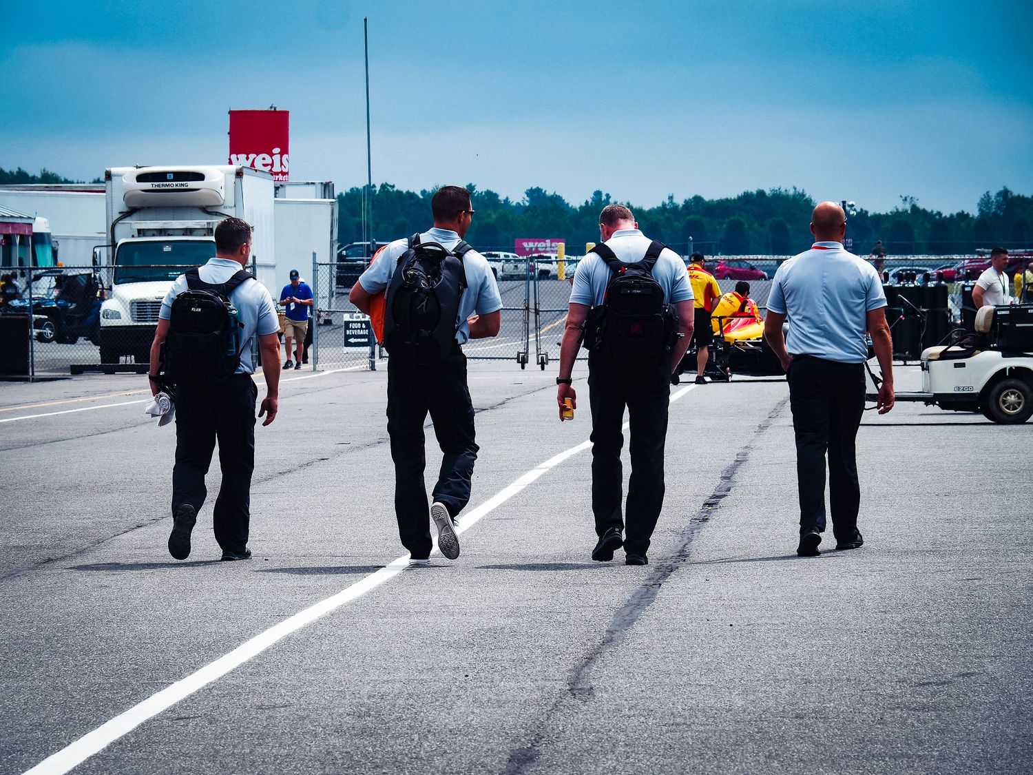 A row of people's legs and feet walking in a line formation on a paved surface against a blue sky background.