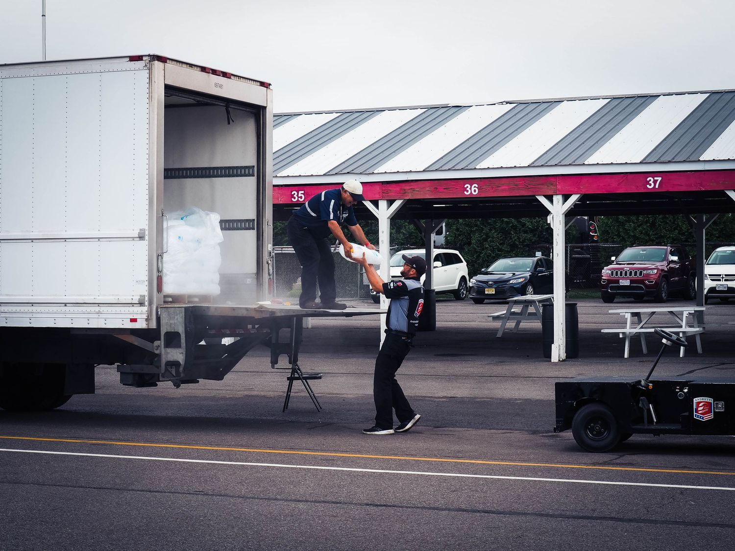 A delivery worker unloads boxes from a truck at a covered parking area on a cloudy day.