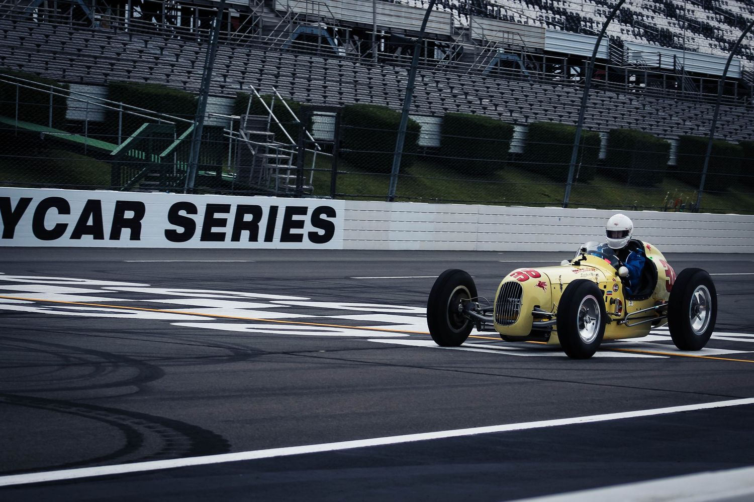 Vintage yellow race car speeds past finish line on professional racing track with empty grandstands in background.