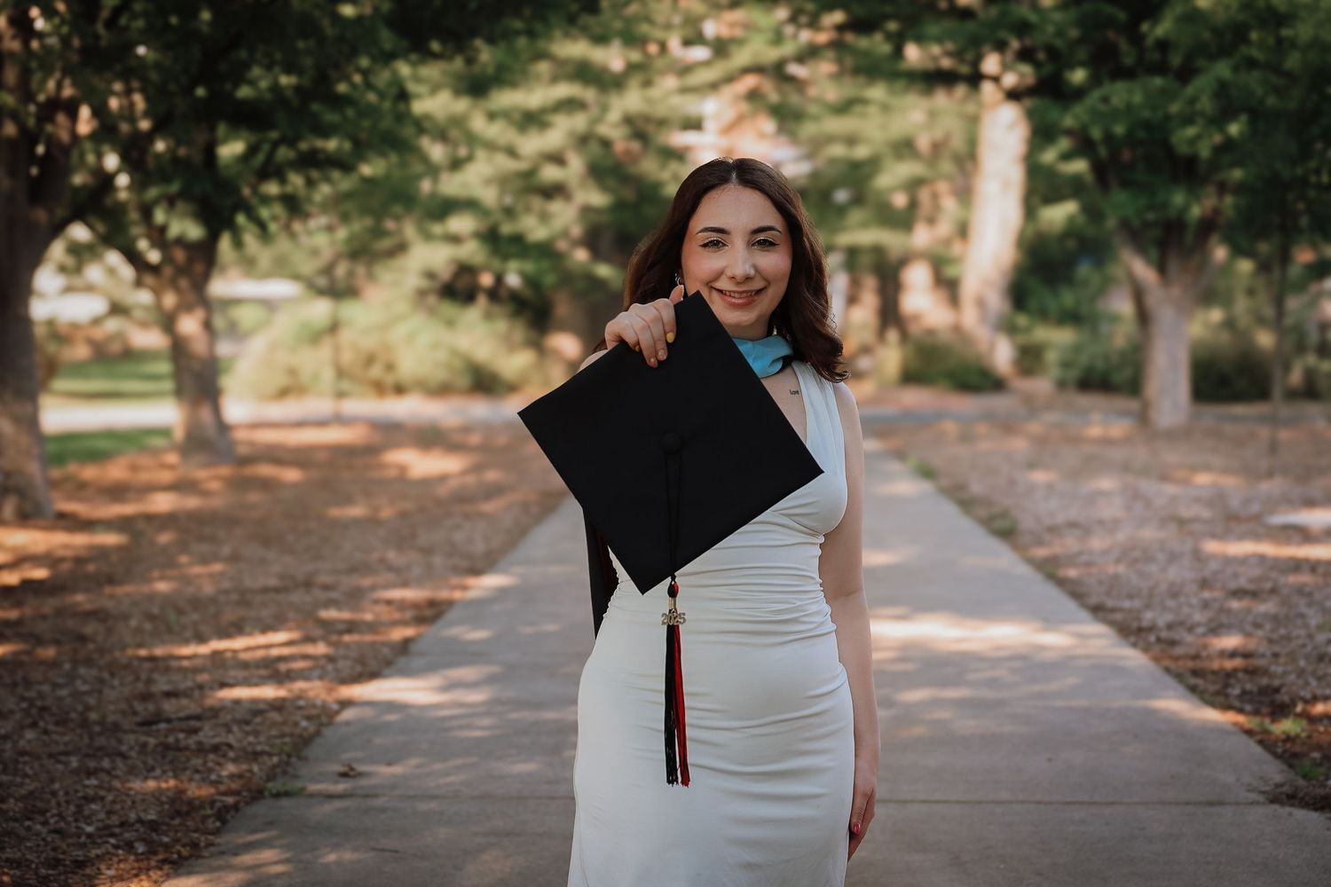 Graduate in white dress celebrates with cap on tree-lined path during outdoor graduation photoshoot.