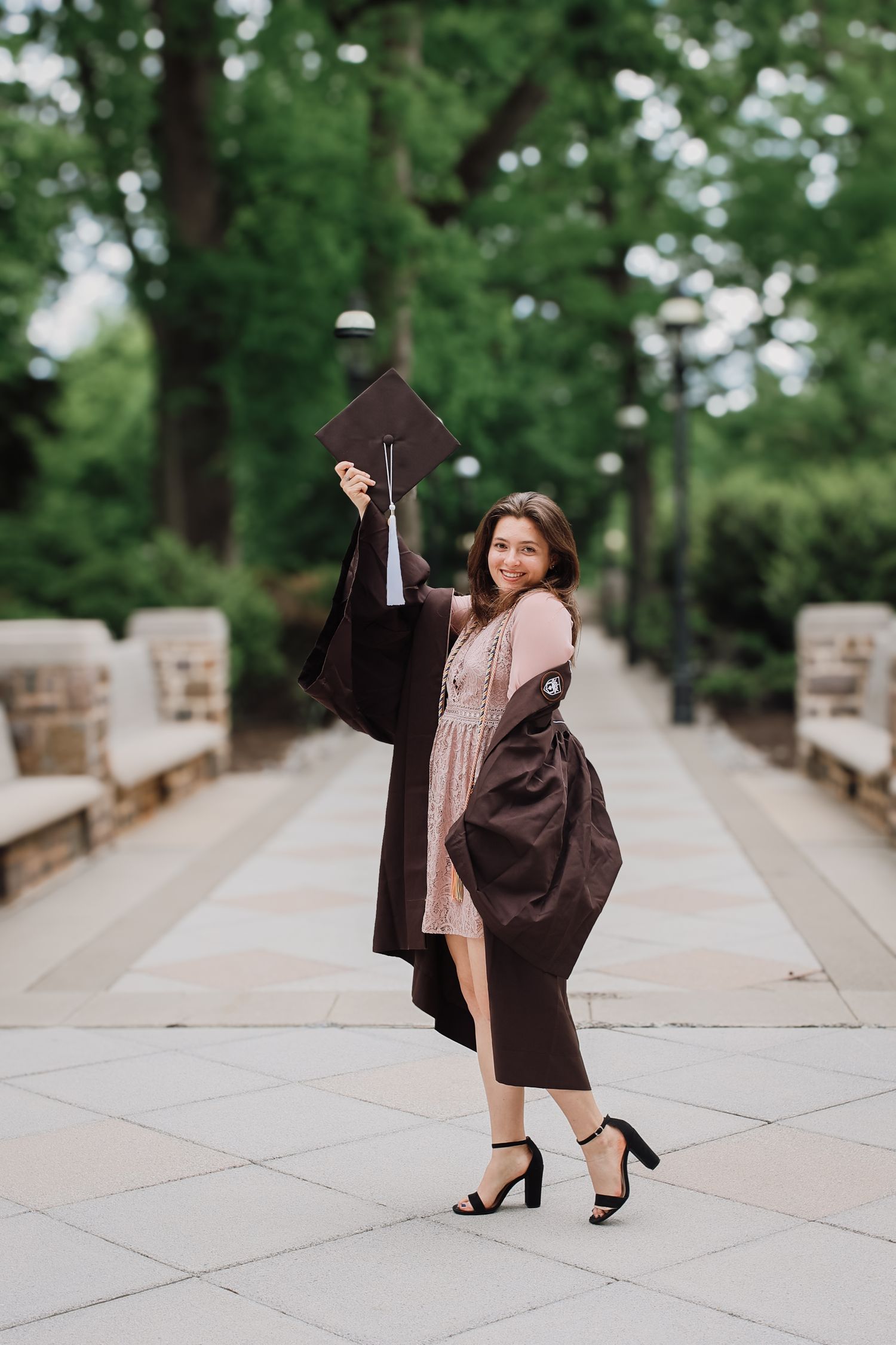 Graduate in brown cap and gown strikes playful poses on concrete walkway during graduation photo session.