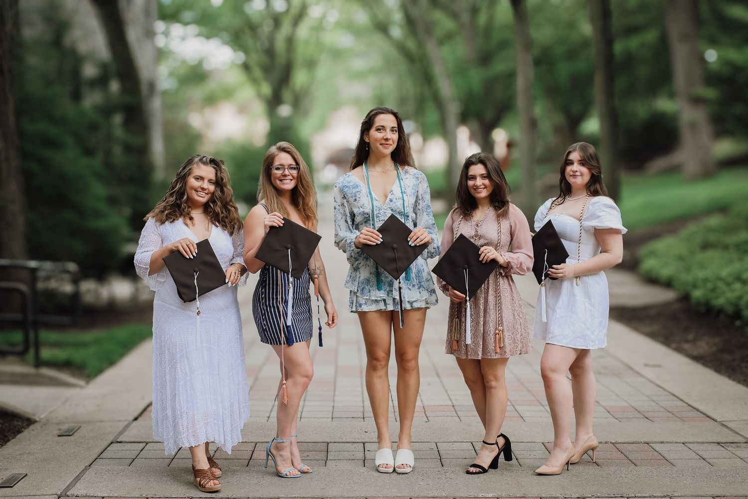A group of friends in casual summer outfits pose together on a stone pathway in a park setting.