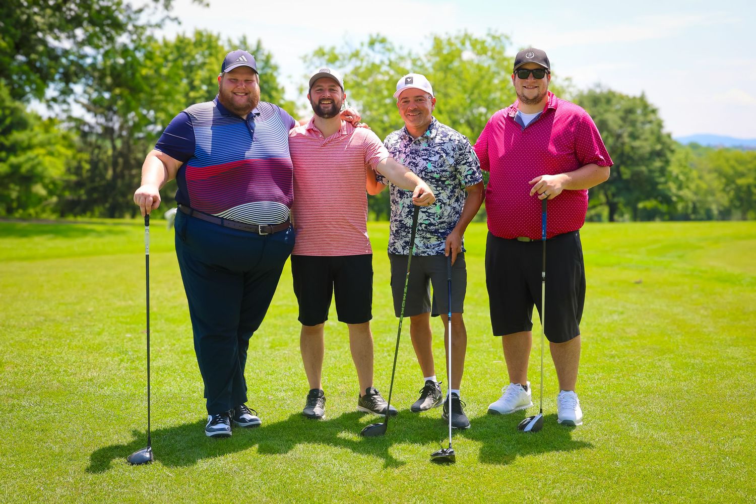 A group of golfers with clubs pose together on a sunny green golf course with trees in the background.