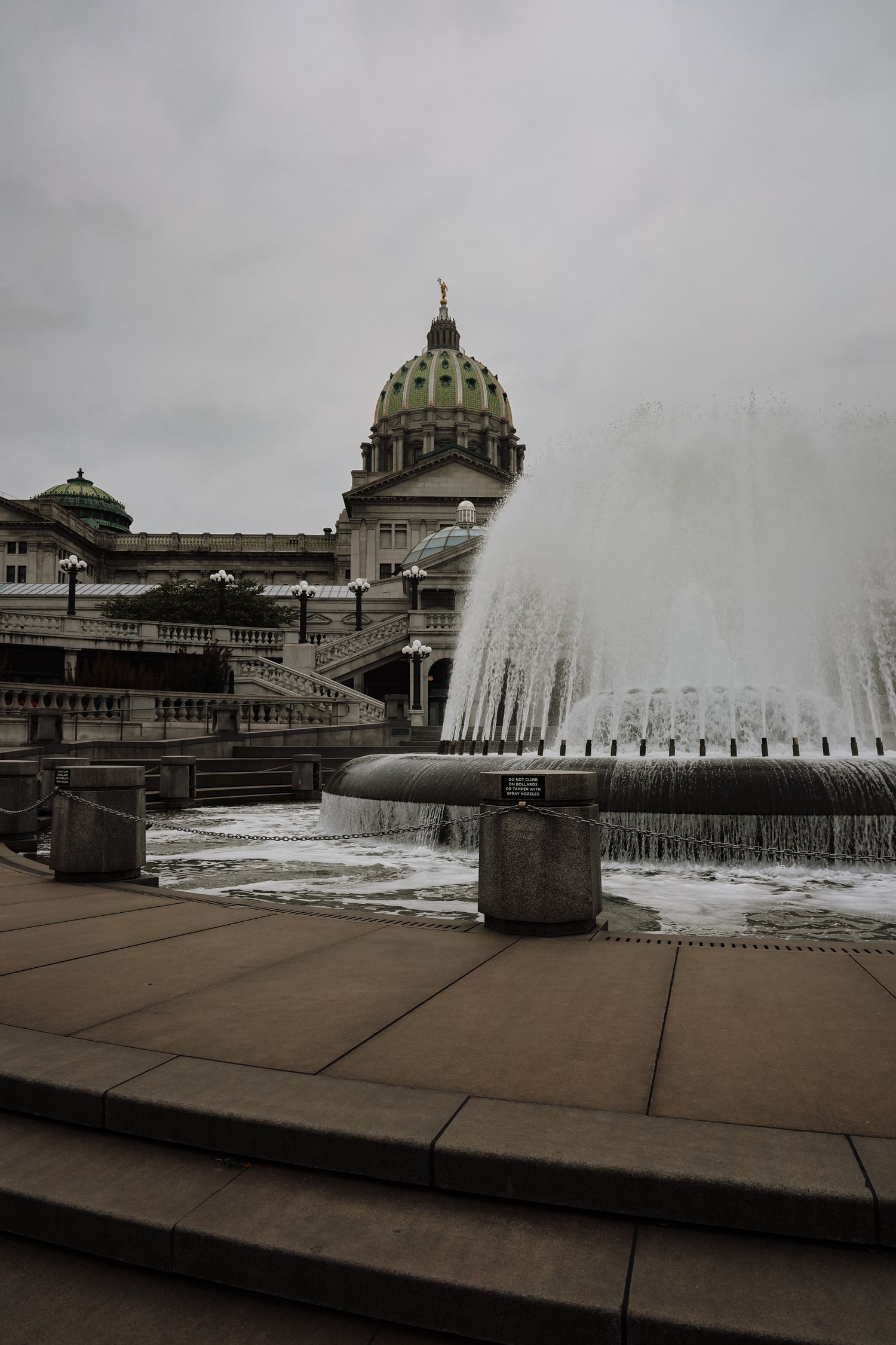 Pennsylvania State Capitol building with green dome behind a large active fountain on an overcast day.