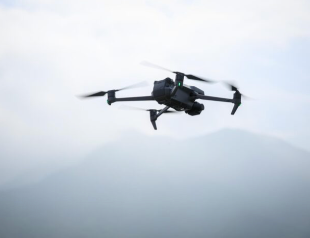 A DJI Mavic drone flies in the sky against a cloudy background with birds visible in the distance.