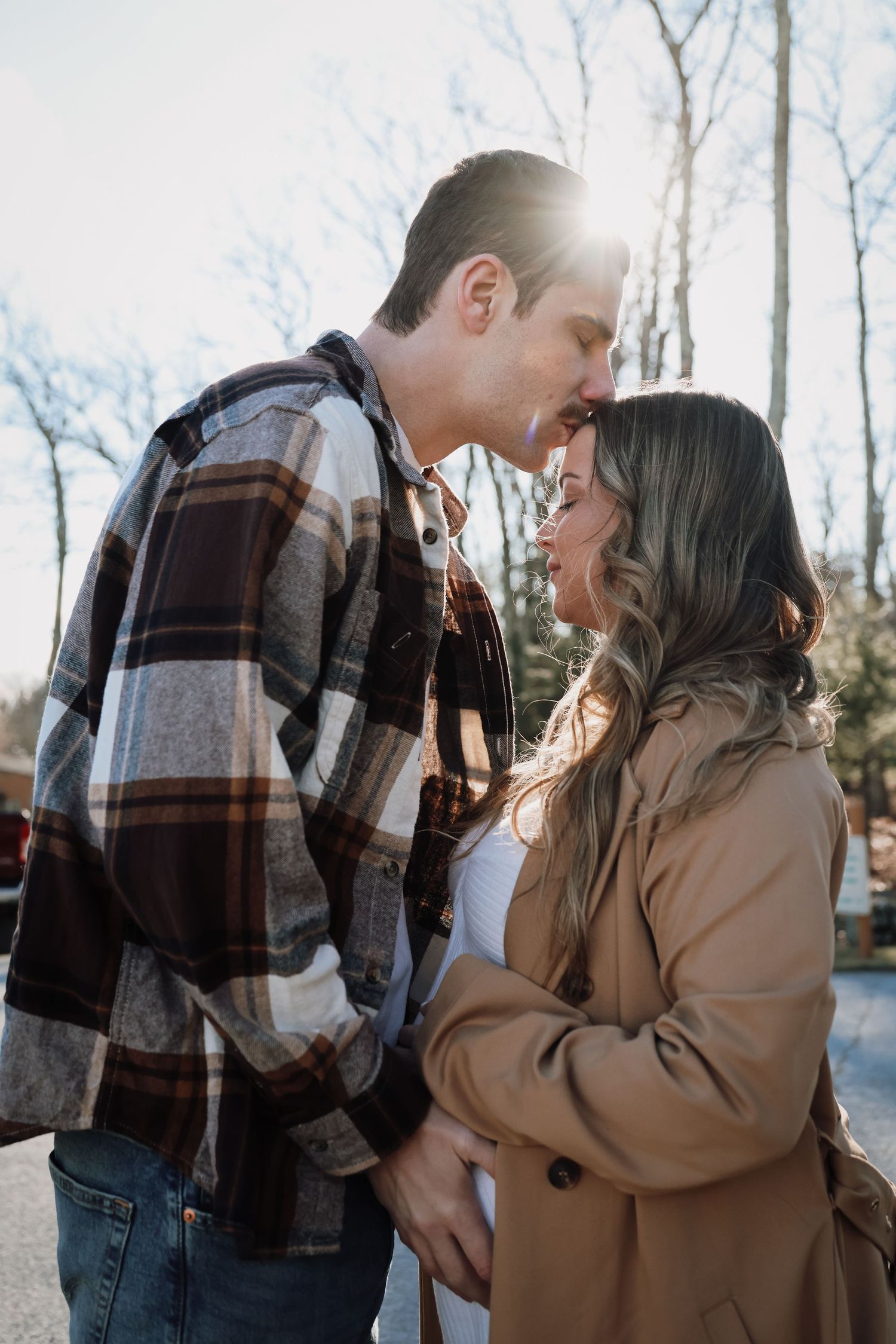 A romantic couple in plaid and tan coats share tender moments in a winter forest setting.