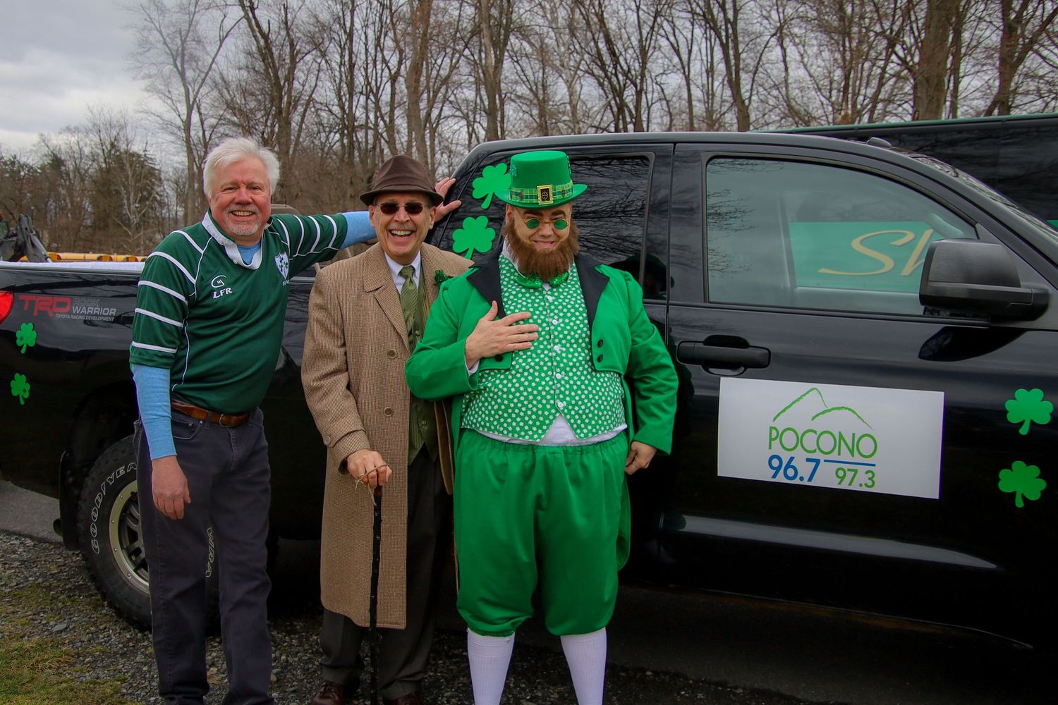 Three people in St. Patrick's Day attire pose next to a black vehicle with a logo and shamrock decoration.