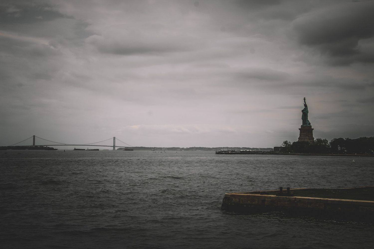 Statue of Liberty standing tall against a dramatic overcast sky with the Verrazano Bridge visible in the background.
