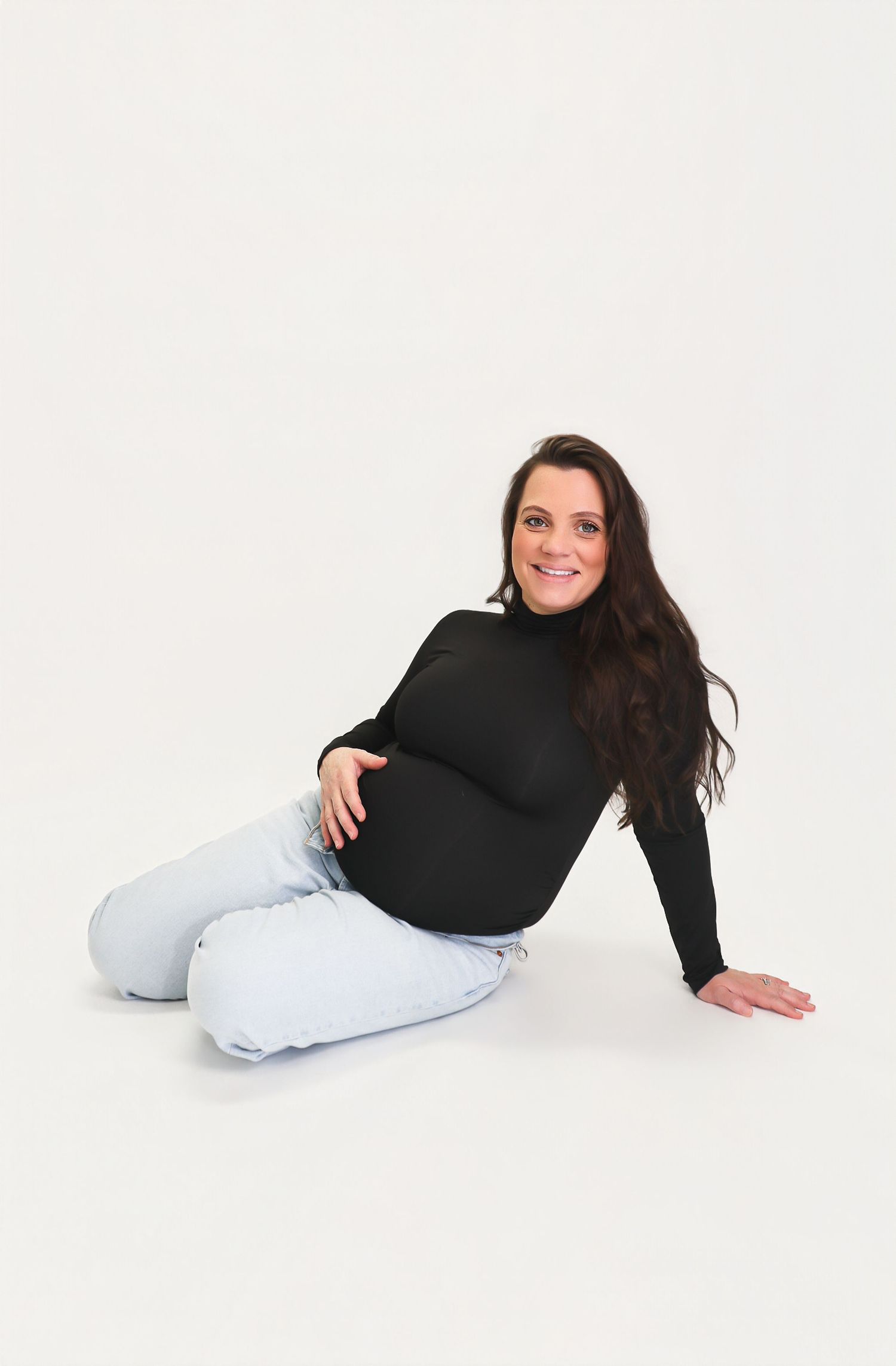 Pregnant woman in black turtleneck and light jeans sitting on floor, smiling, showcasing maternity fashion.