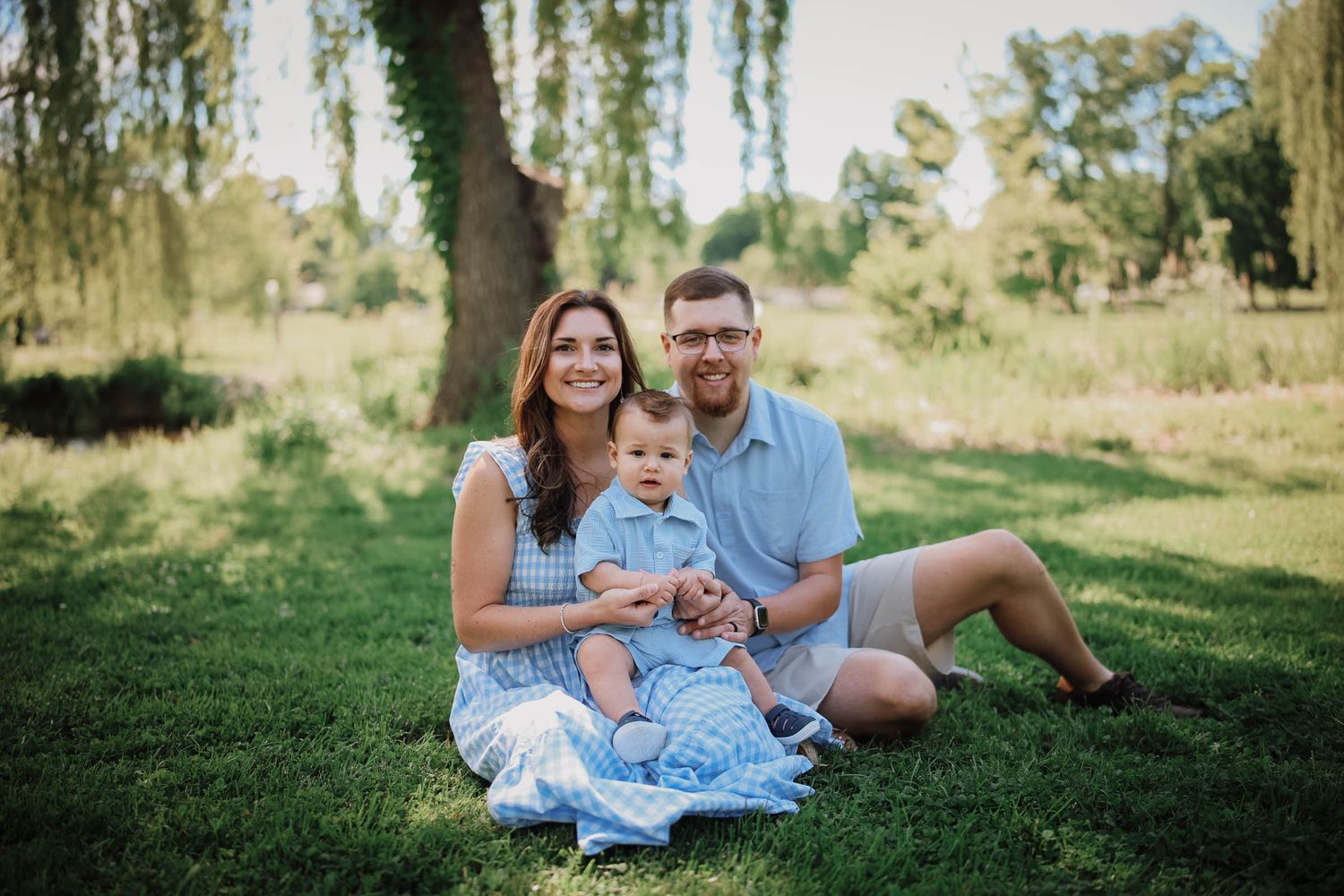A happy young family with a baby sits on green grass in a sunny park with willow trees in the background.