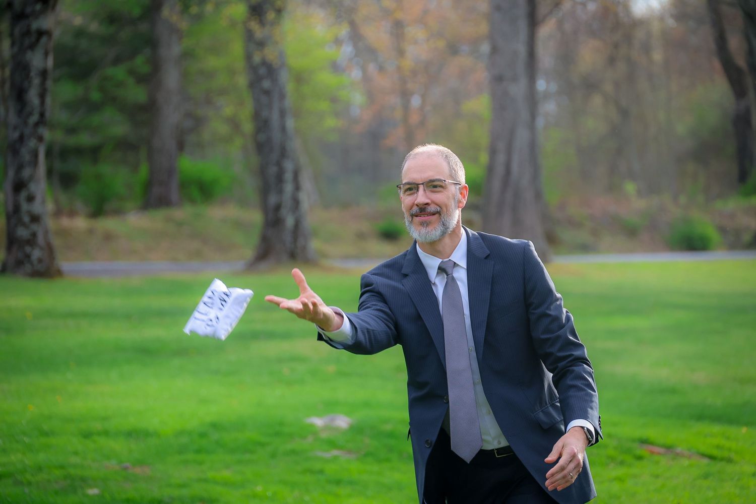 Smiling businessman in suit tossing paper outdoors in lush green park surrounded by trees.