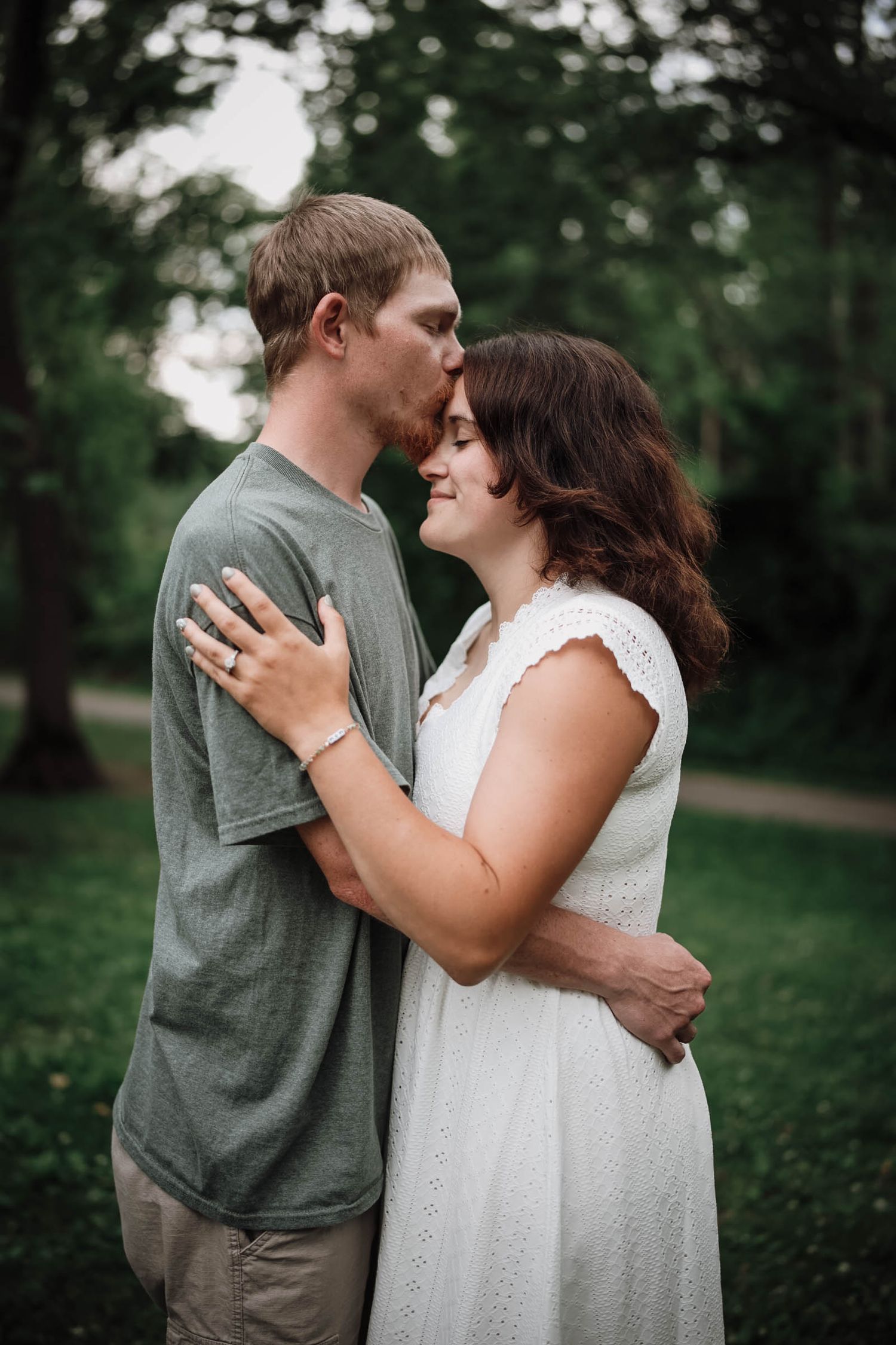 A couple embraces outdoors, man kissing woman's forehead, she smiles in white dress, green trees in background.