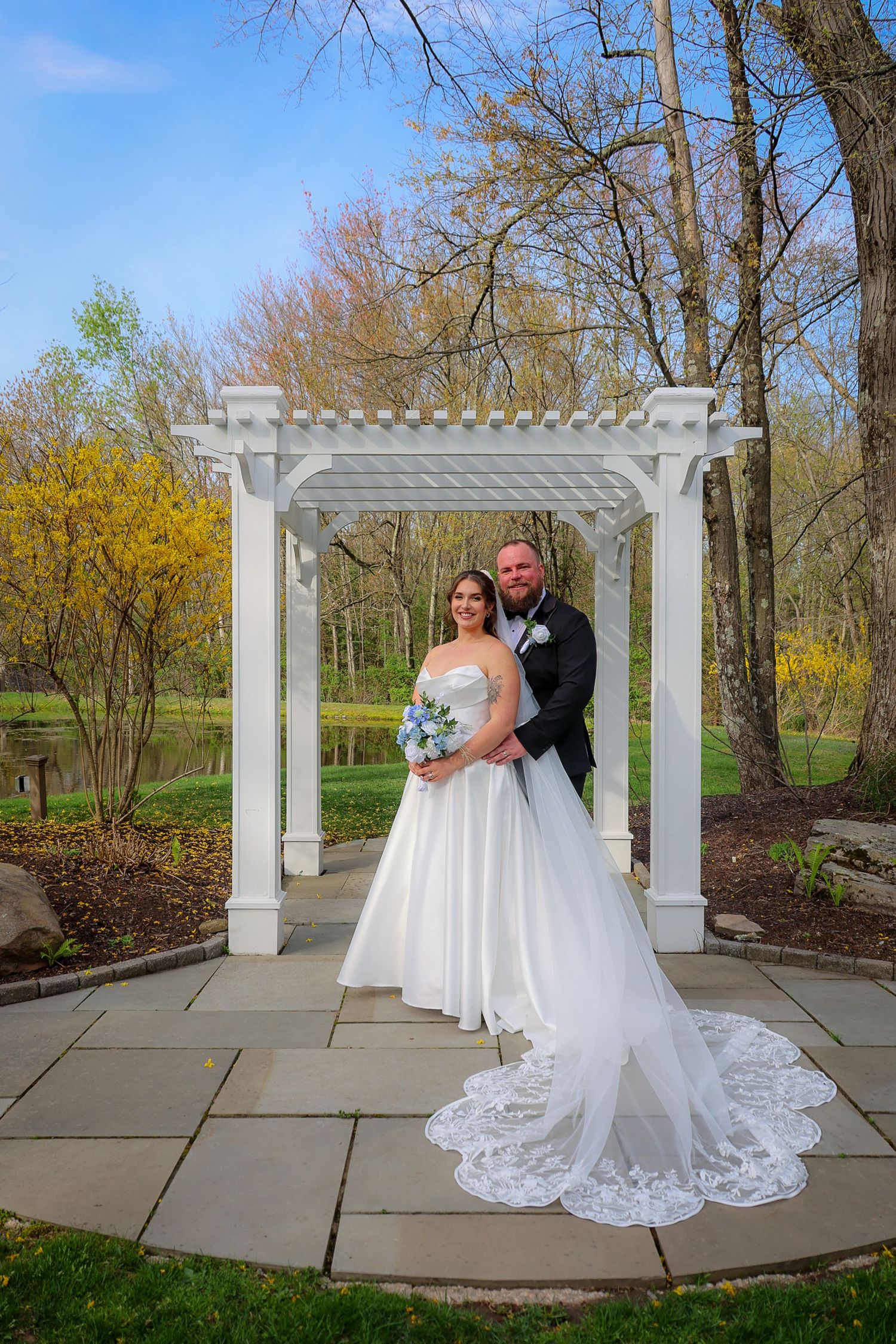 Bride and groom pose under white pergola in lush garden setting, bride in elegant gown with lace train holding blue bouquet.