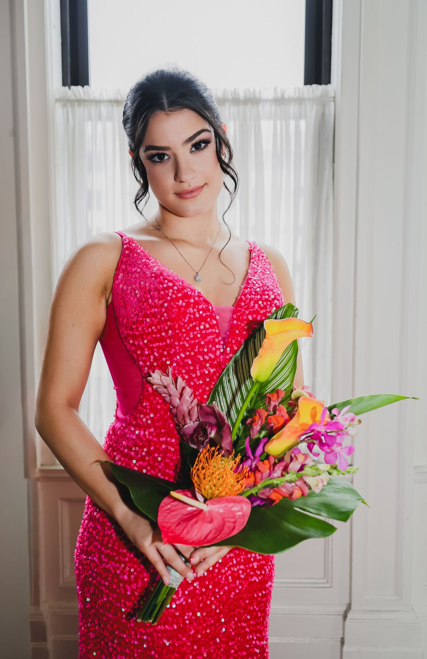 Young woman in a pink sequin gown holding a vibrant tropical bouquet with calla lilies and anthuriums near a window.