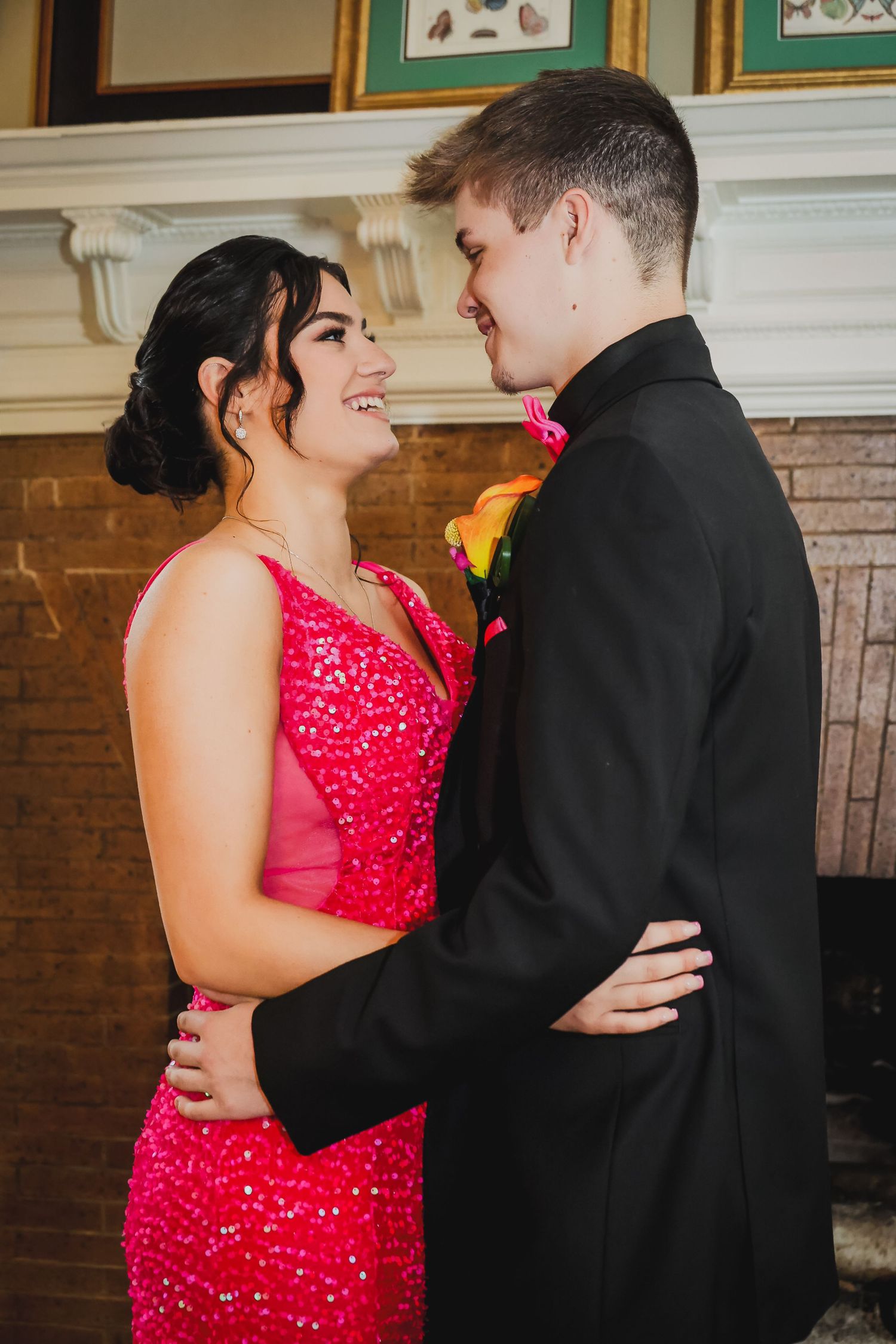 A smiling couple in prom attire, girl in pink sequin dress and boy in black suit with floral boutonniere.