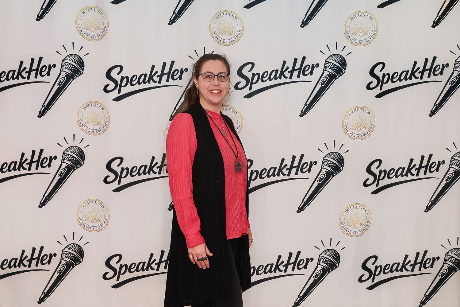 Woman in red blazer poses at SpeakHer event backdrop with microphone logos and branding.