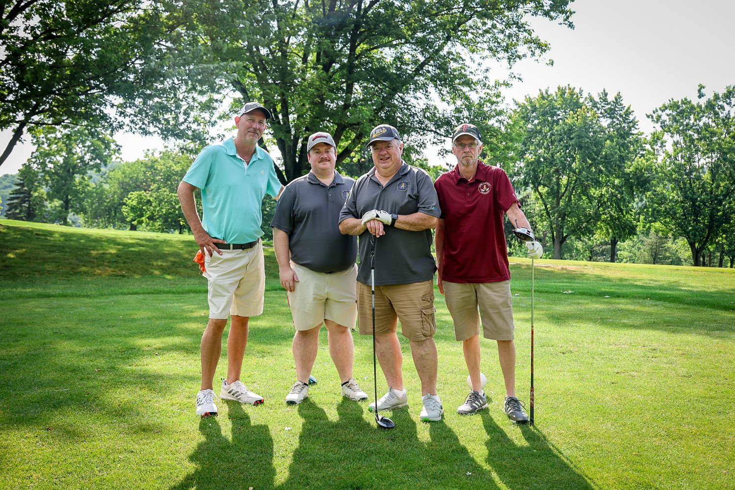 Four men posing together on a sunny golf course, holding clubs and smiling during a golf outing.