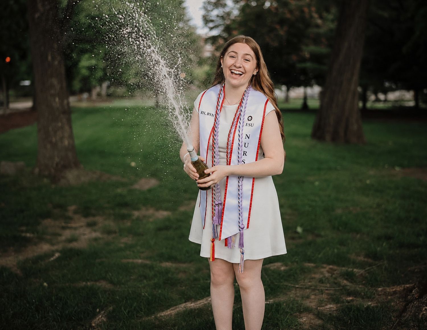 Smiling female graduate in white dress and honor stoles celebrates outdoors by spraying champagne.