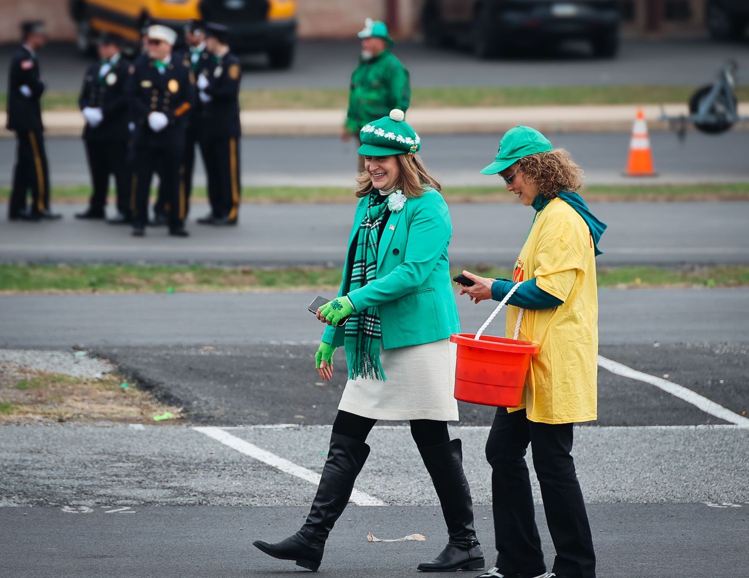 Two women in green St. Patrick's Day attire walk in a parking lot during a parade, with uniformed officers in the background.