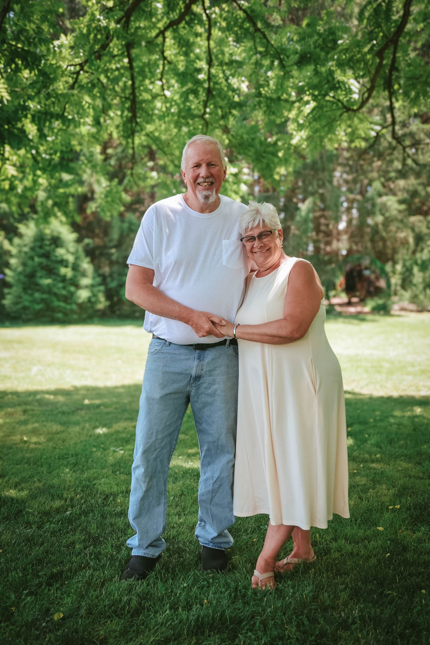 A smiling older couple holds hands while standing together on a green lawn surrounded by trees.