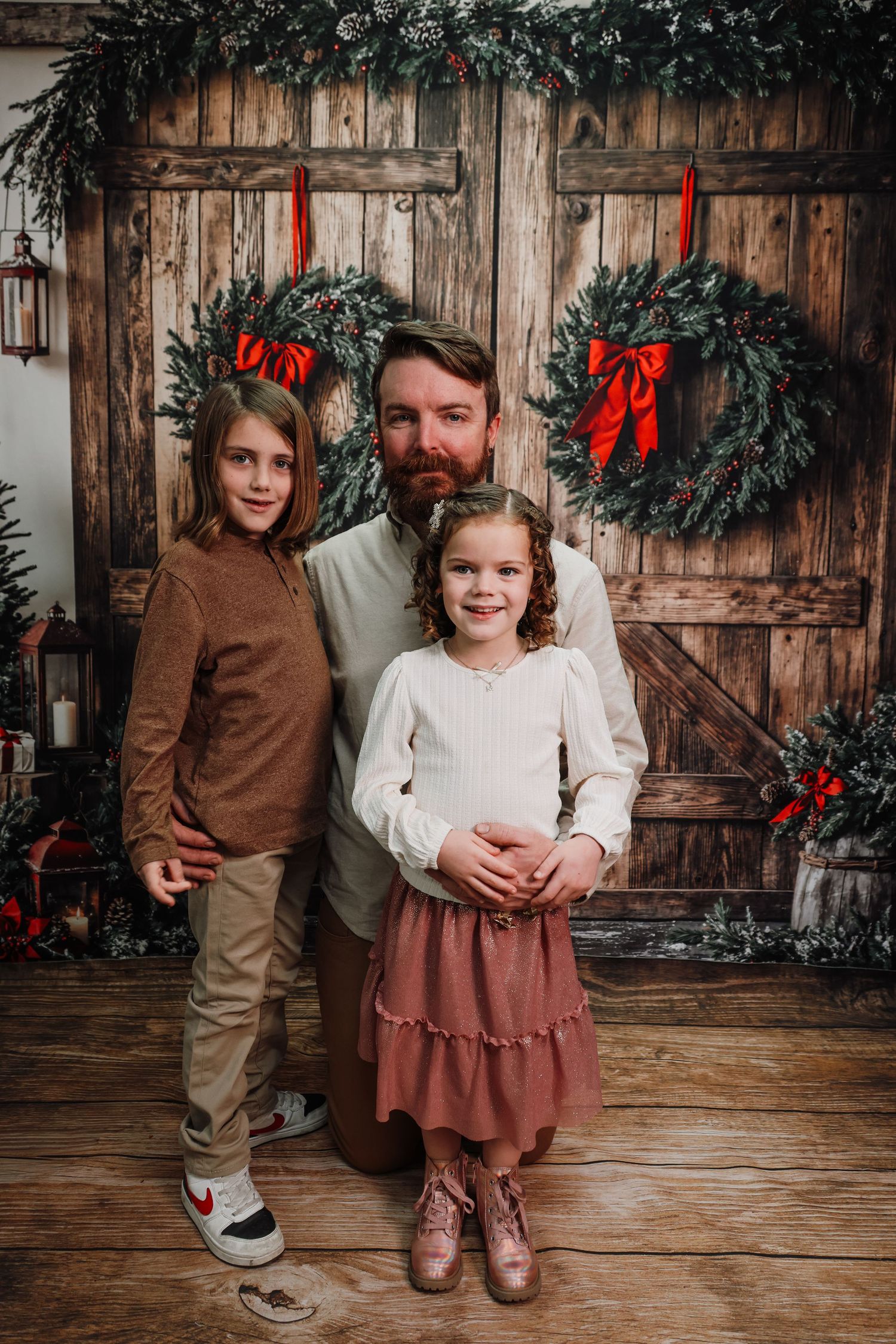 A man and two young girls pose for a Christmas family portrait in front of rustic wooden doors with holiday wreaths.