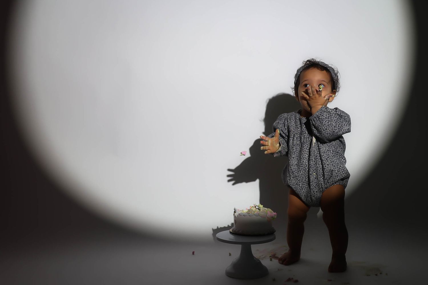 Toddler in gray romper stands by smashed birthday cake, covering mouth with hand in surprise.