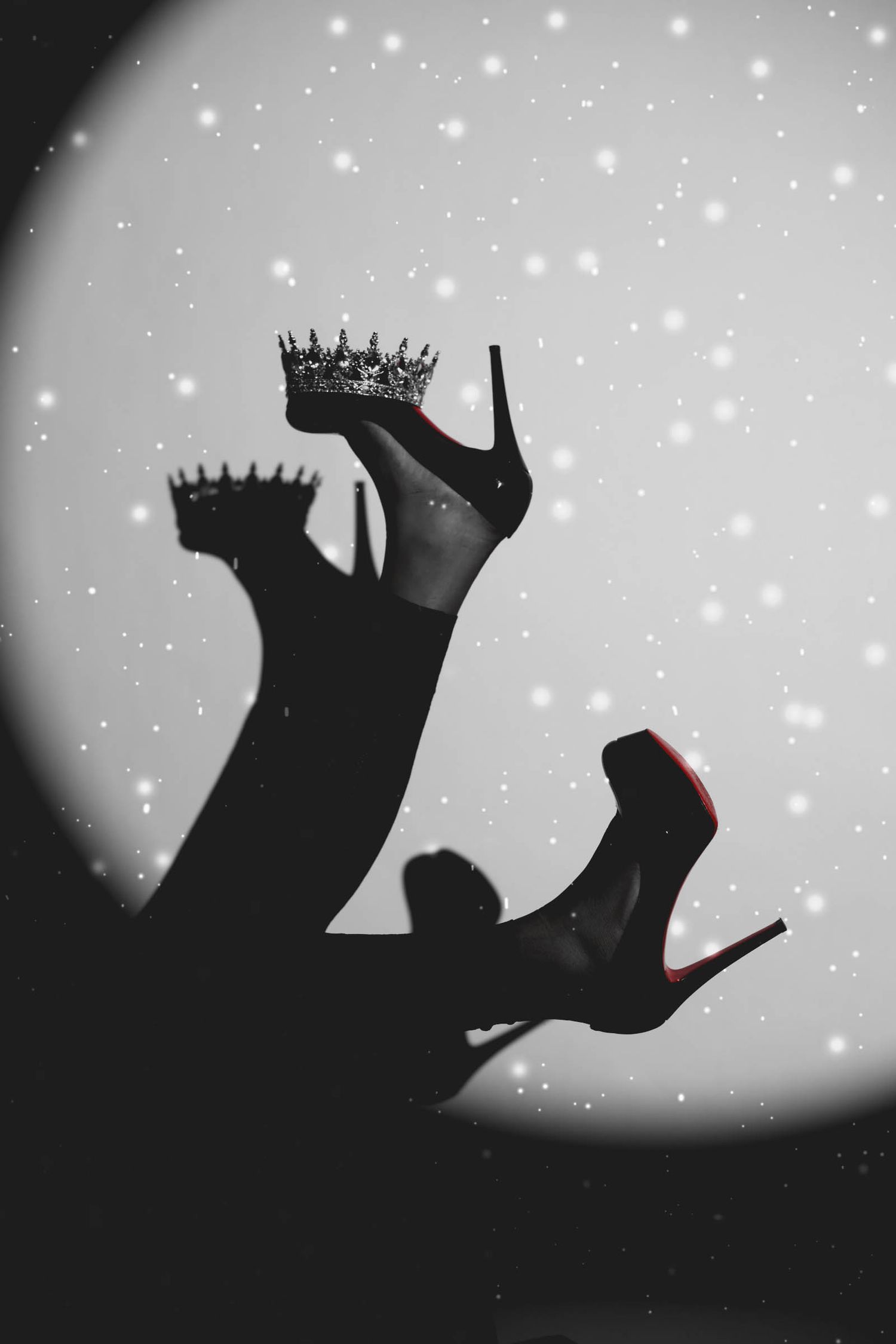Black and white artistic photo of high heels with red soles and crown balanced on heel against glittery spotlight.