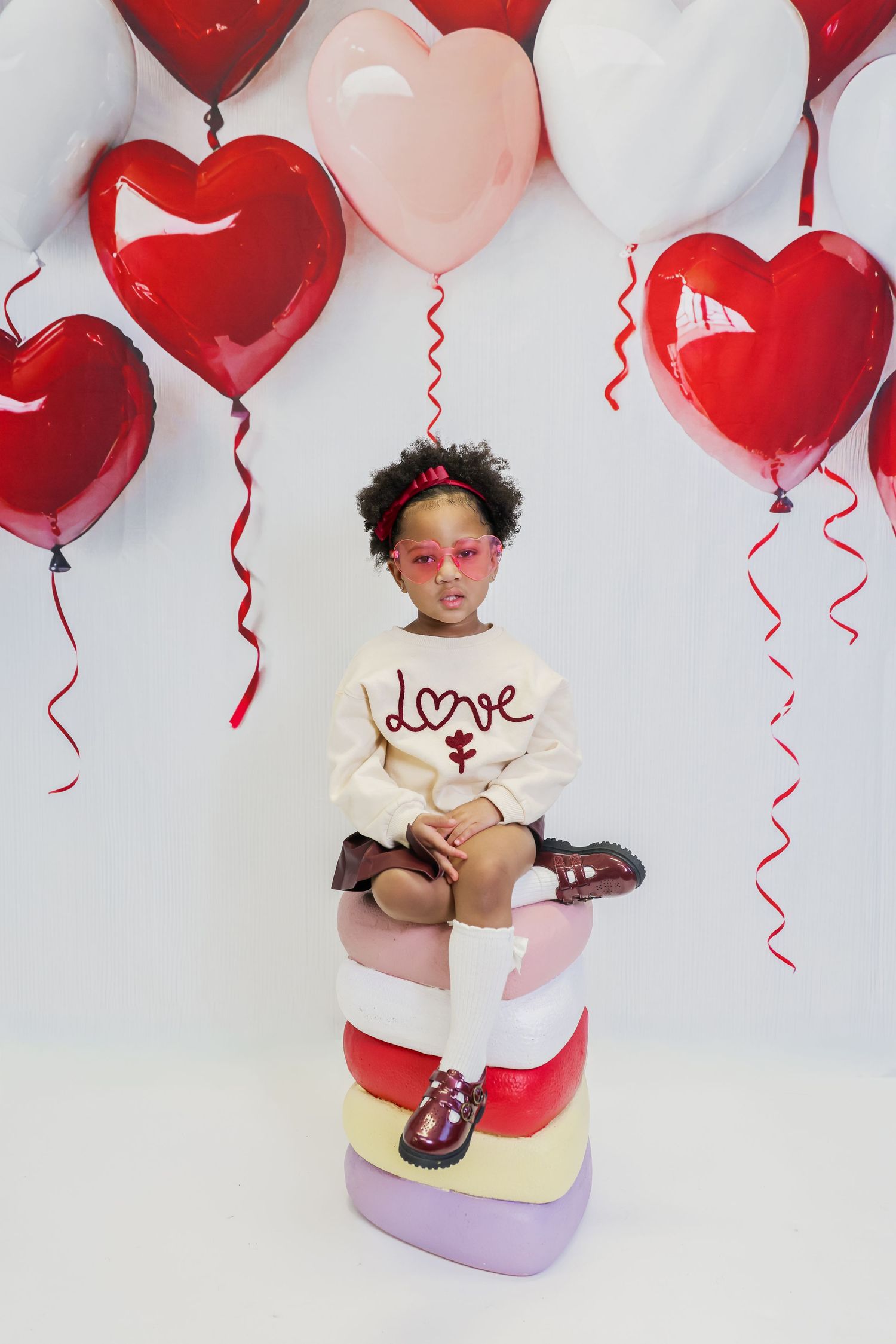 A young girl in a cream ‘Love’ sweater and pink heart sunglasses sits on stacked hearts surrounded by red and pink heart balloons.
