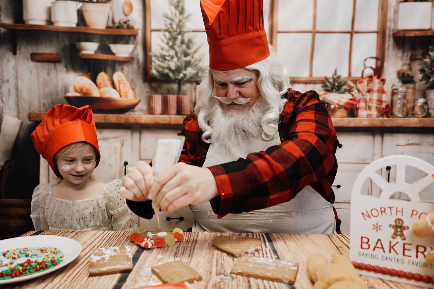 Santa Claus and child in red chef hats decorating Christmas cookies at North Pole Bakery kitchen table.