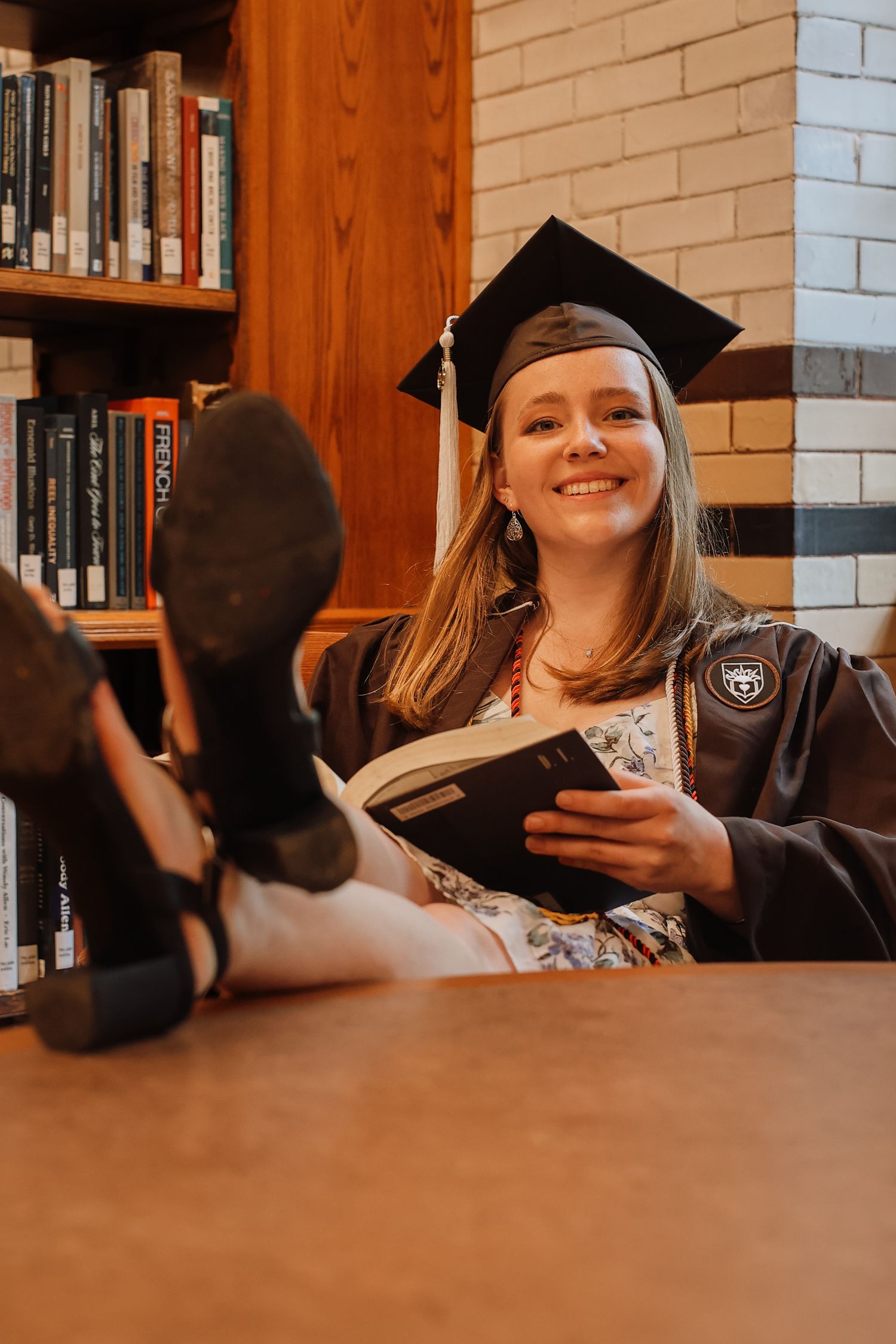 A graduate in cap and gown poses with books in a library setting with warm lighting and wooden shelves.
