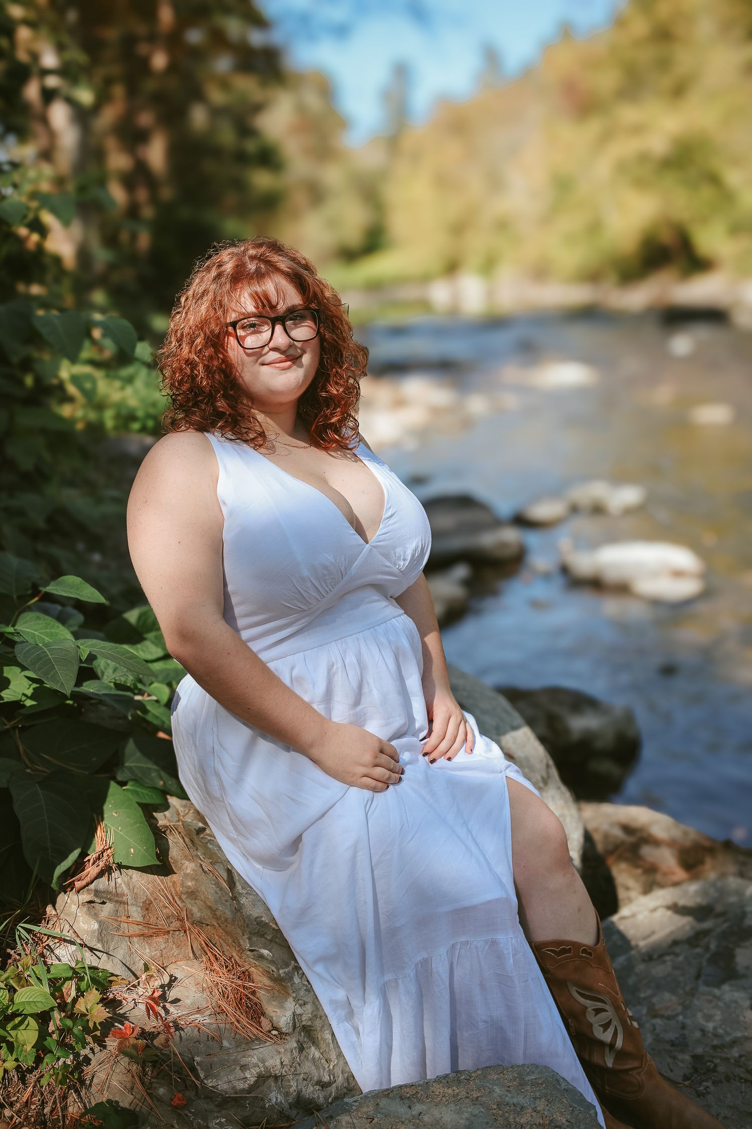 Woman in white dress sits gracefully on rocks by a flowing stream on a sunny day.