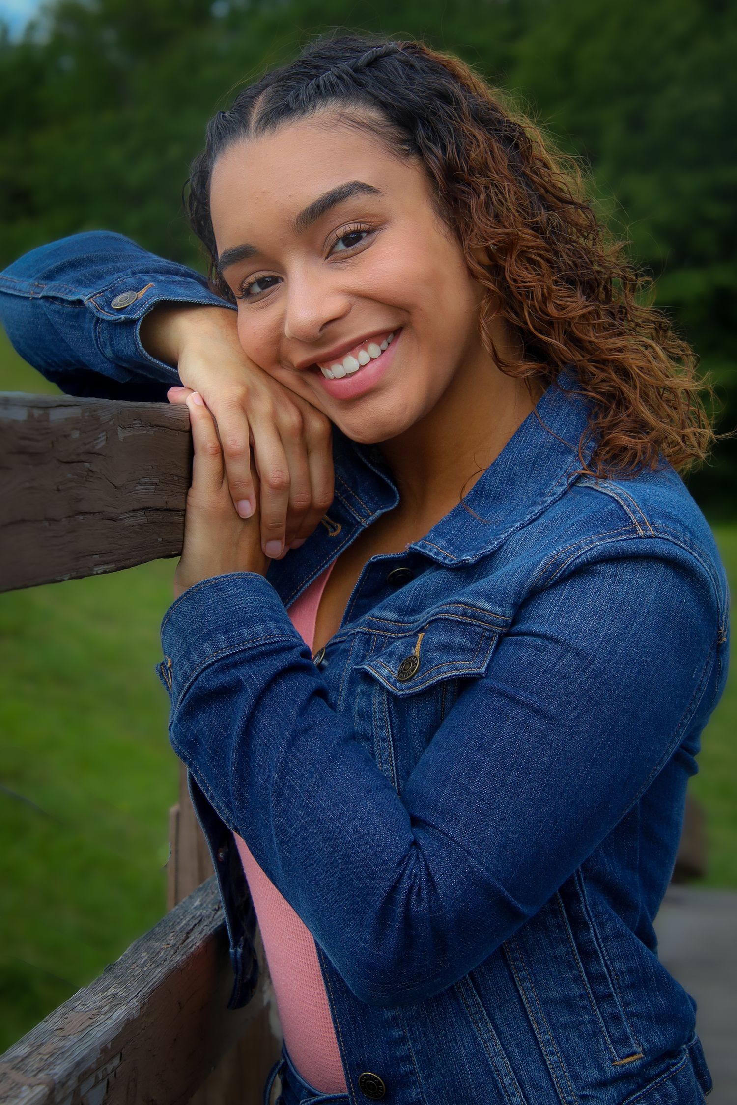 A cheerful young woman in a denim jacket leans on a wooden railing outdoors with natural greenery in the background.