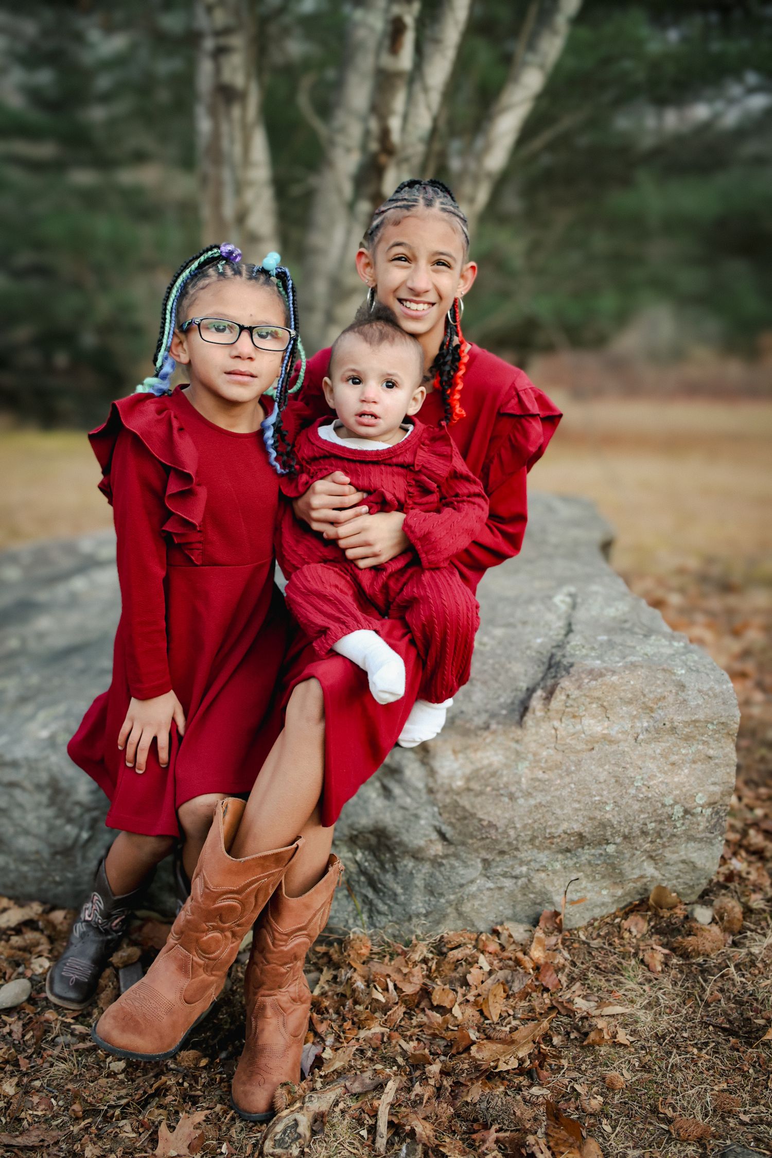 Two children in matching red coats sit together on large rocks surrounded by fallen autumn leaves.