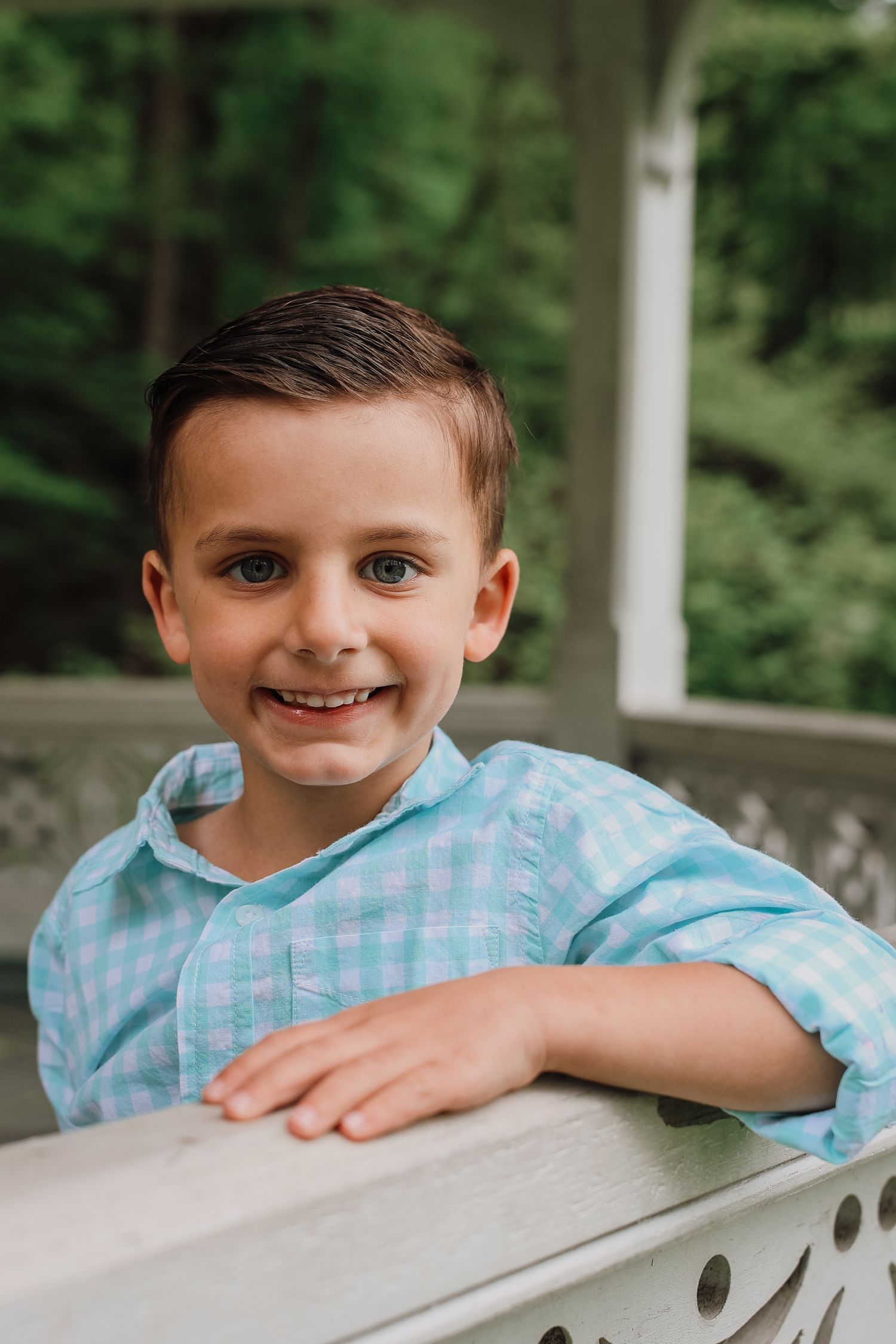 A child in a light blue checkered shirt leans on a white railing outdoors with trees in the background.