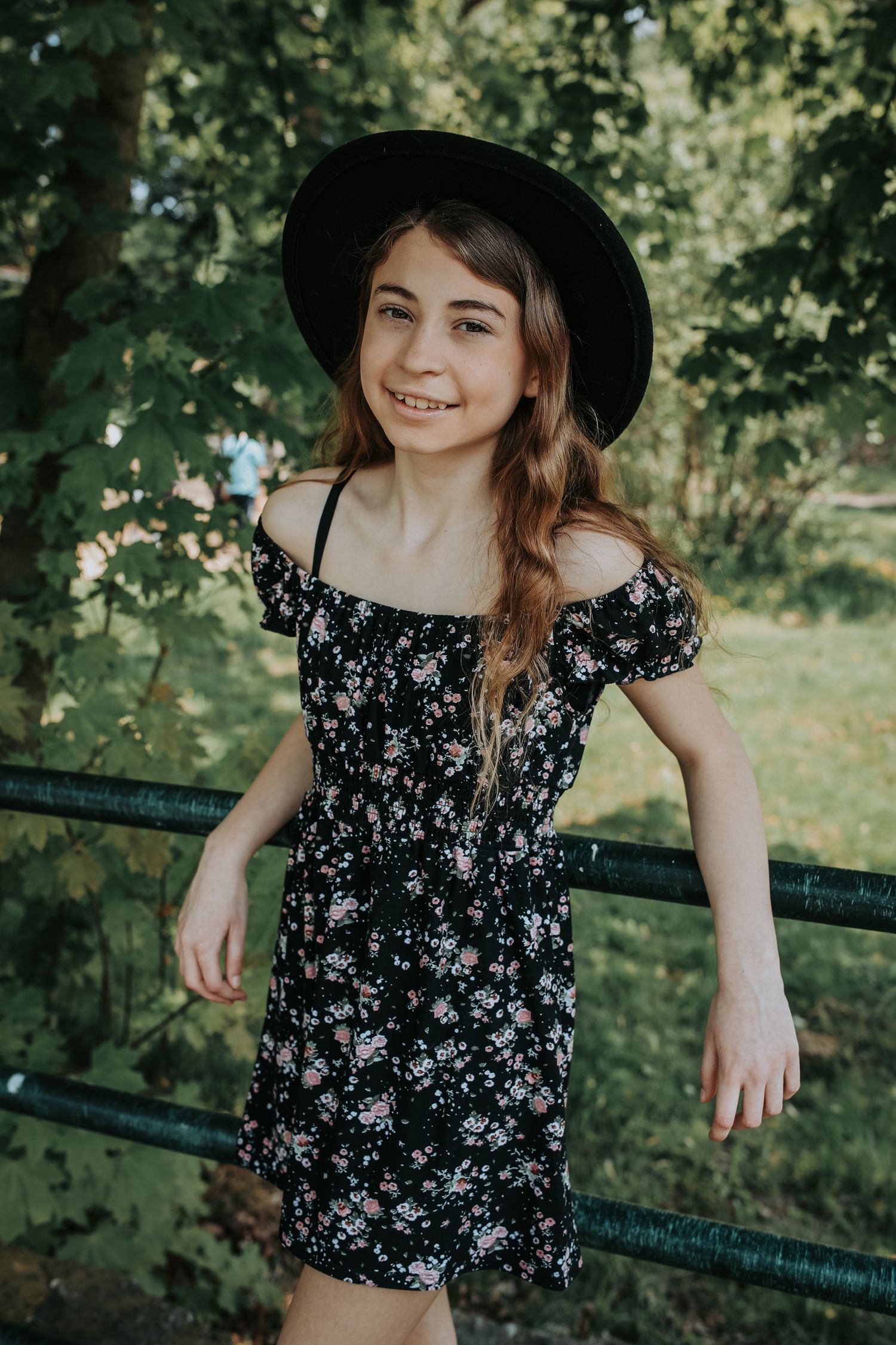 A person in a black floral dress and hat poses playfully in a park setting with lush green trees in the background.