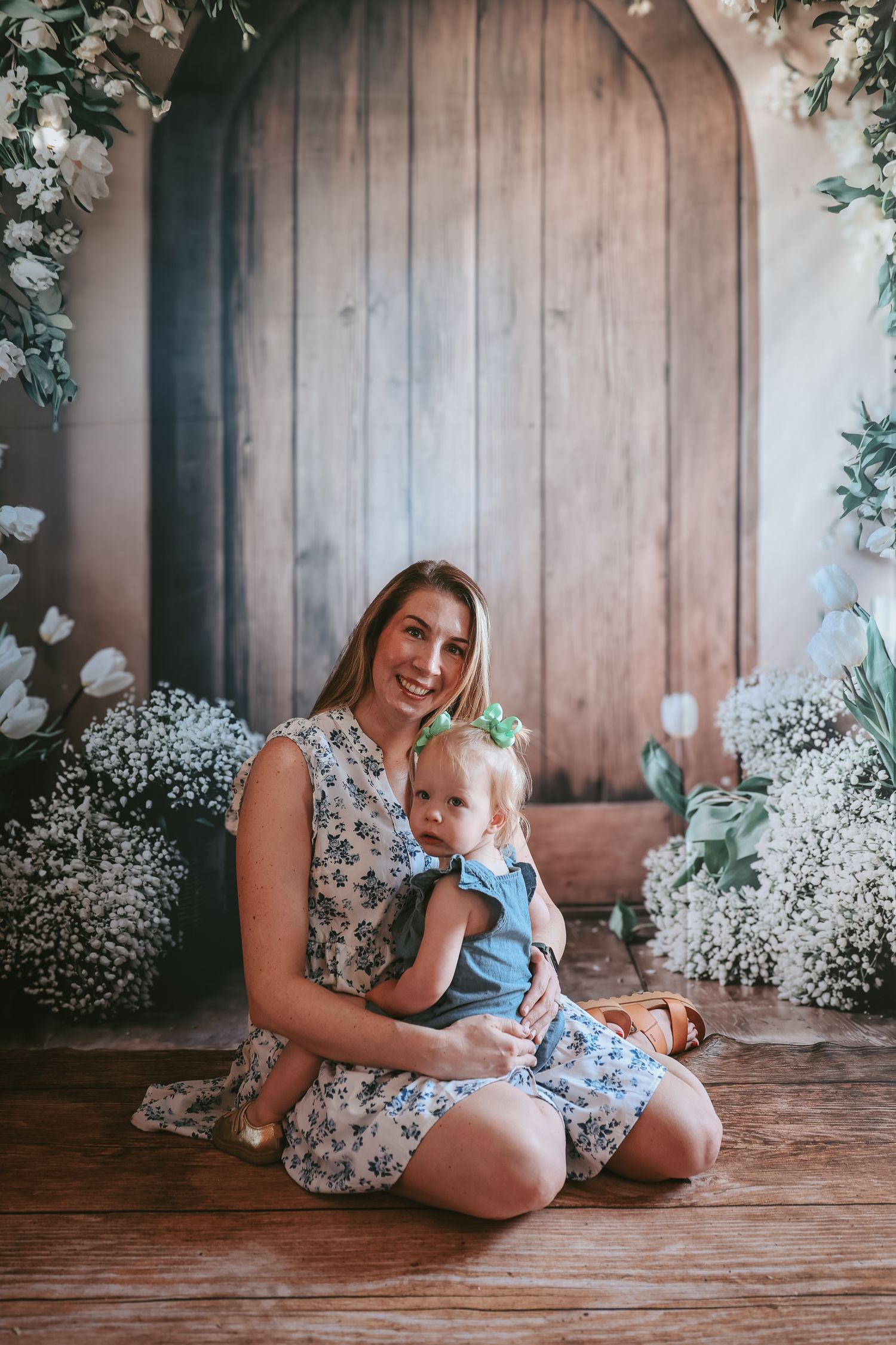 A mother and baby share tender moments on a rustic wooden floor surrounded by delicate white baby's breath flowers.
