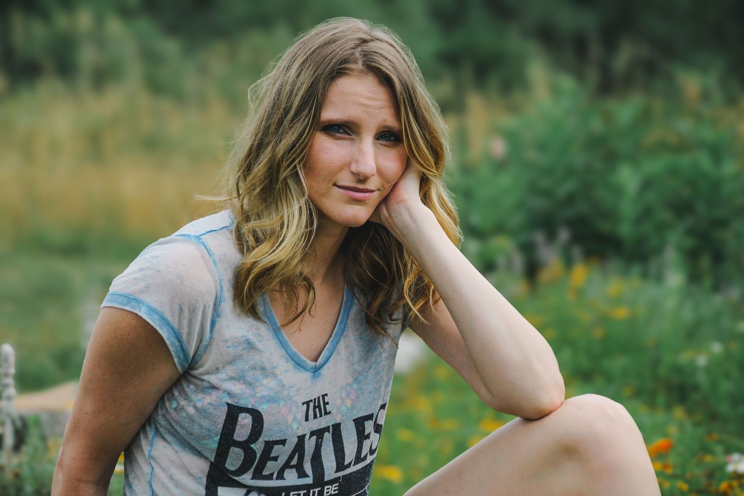 A person in a Beatles t-shirt poses outdoors in a field with yellow wildflowers and green foliage.