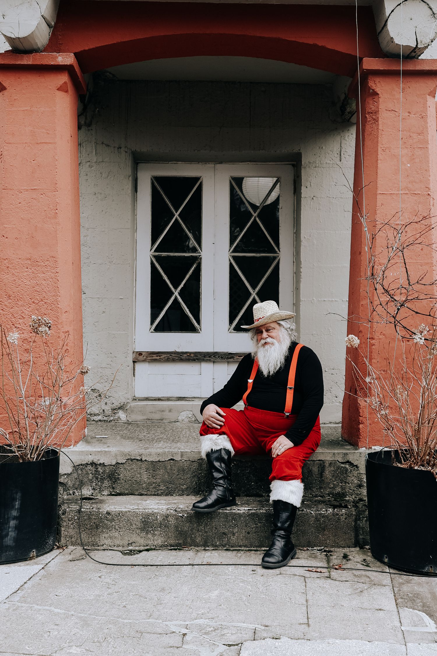 A figure in a red coat and black boots sits on a concrete ledge outside a building with white-framed windows.