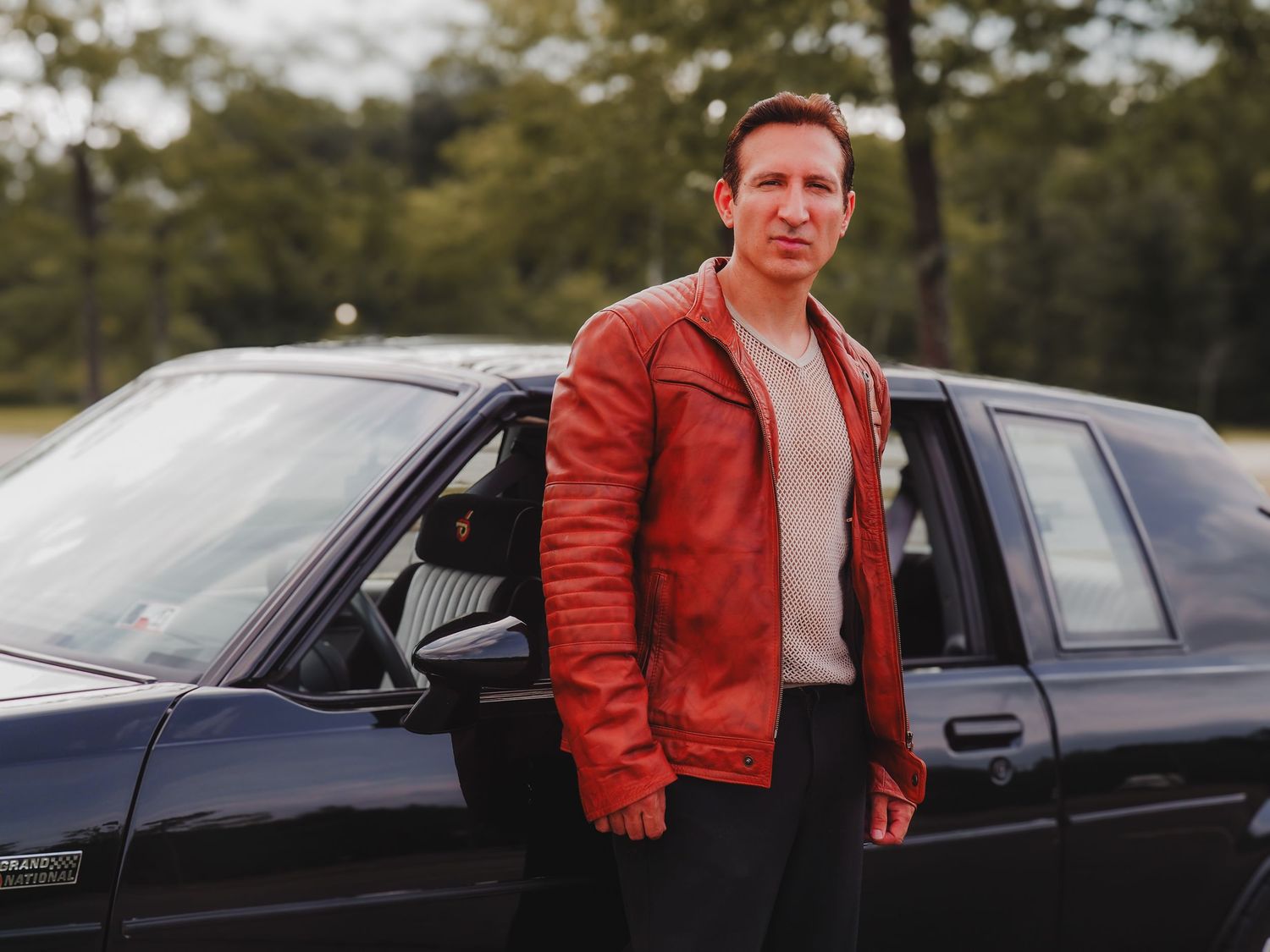 Person in red leather jacket poses next to black car in outdoor setting with trees in background.