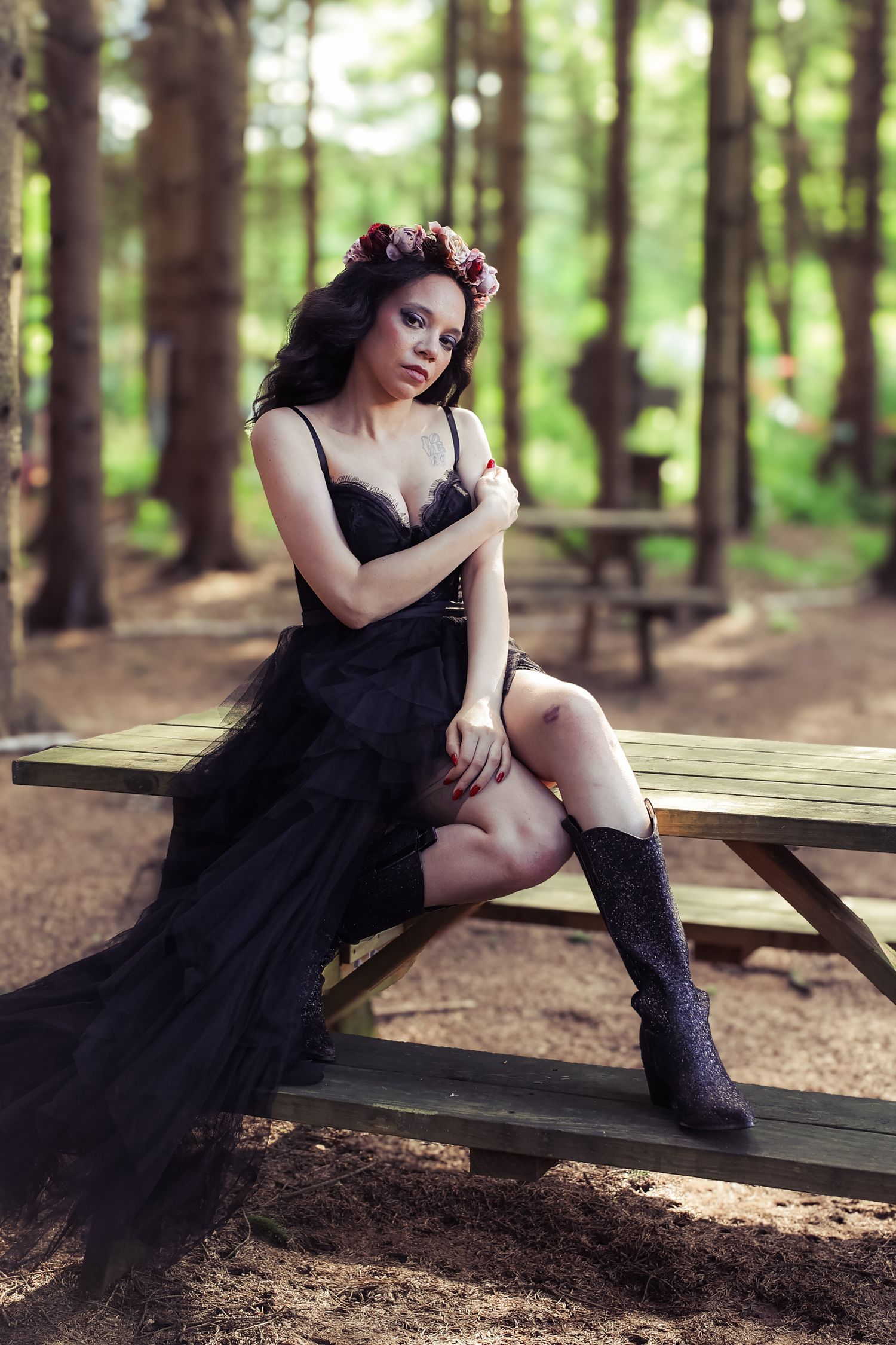 A figure in a black dress and boots poses on a wooden picnic table in a serene forest setting.